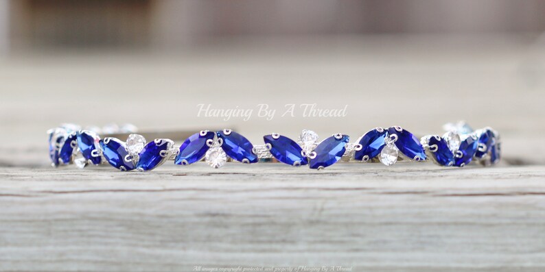 Pu&ograve; includere: Un braccialetto d'argento con strass a forma di marquise blu e trasparenti alternati. Il braccialetto &egrave; su una superficie di legno.
