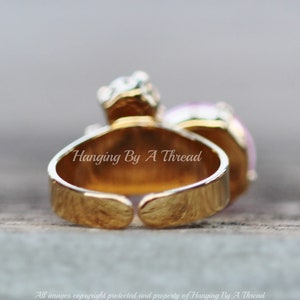 May include: A gold-toned adjustable ring with a large, light purple gemstone and smaller clear stones. The ring has the text "Hanging By A Thread" on the band. The ring is set against a blurred background.