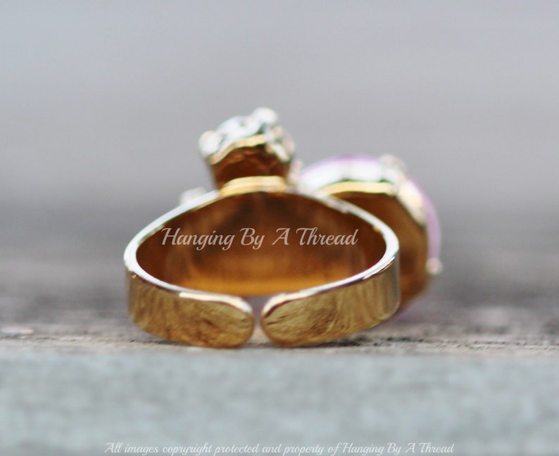 May include: A gold-toned adjustable ring with a large, light purple gemstone and smaller clear stones. The ring has the text "Hanging By A Thread" on the band. The ring is set against a blurred background.