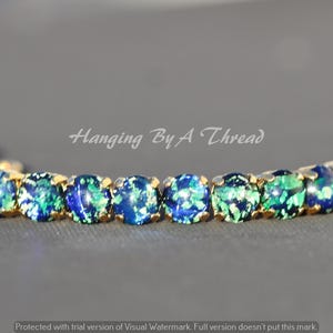 May include: A gold-tone bracelet featuring a row of round, iridescent blue and green stones. The text "Hanging By A Thread" is visible above the bracelet. The stones exhibit a speckled, reflective appearance with deep blue and bright green hues.