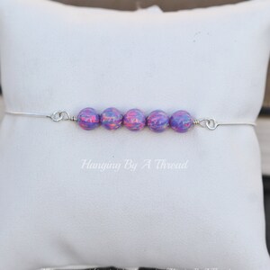 Puede incluir: Una pulsera de plata con una fila de seis cuentas iridiscentes de color p&uacute;rpura y rosa. La pulsera est&aacute; sobre un fondo blanco.