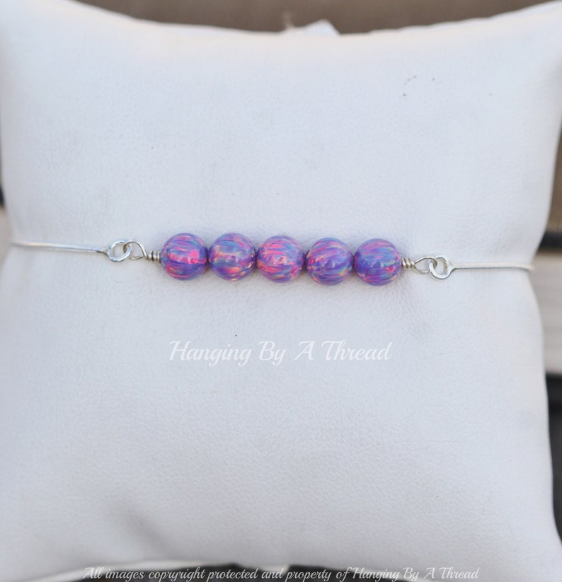 Puede incluir: Una pulsera de plata con una fila de seis cuentas iridiscentes de color p&uacute;rpura y rosa. La pulsera est&aacute; sobre un fondo blanco.