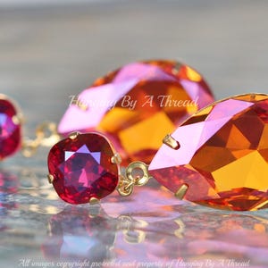 May include: A pair of dangle earrings featuring a teardrop-shaped orange and pink crystal and a square red crystal. The earrings are made with gold-toned metal.
