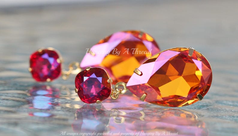 May include: A pair of dangle earrings featuring a teardrop-shaped orange and pink crystal and a square red crystal. The earrings are made with gold-toned metal.