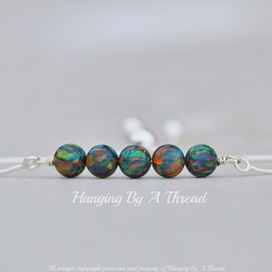 May include: A silver bangle bracelet with five iridescent green, blue, and orange beads. The bracelet is open and has a simple design. "Hanging By A Thread" is written below the bracelet.