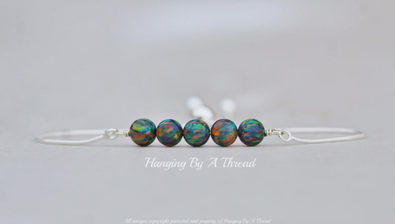 May include: A silver bangle bracelet with five iridescent green, blue, and orange beads. The bracelet is open and has a simple design. "Hanging By A Thread" is written below the bracelet.