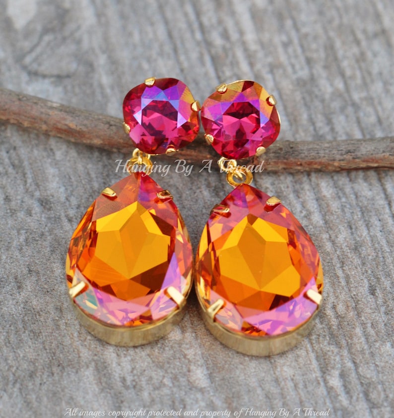 May include: Gold dangle earrings featuring a teardrop-shaped orange and pink iridescent crystal, with a smaller square pink iridescent crystal.