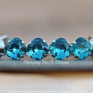 以下が含まれることがあります： 銀色の枠にセットされた、7つのターコイズブルーの丸いラインストーンが並んでいます。ラインストーンは灰色の表面に一直線に並んでいます。ラインストーンの下には「Hanging By A Thread」と書かれています。