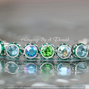 May include: A silver bracelet with a series of round, iridescent gemstones in shades of green and blue. The bracelet is set against a blurred background, with the text "Hanging By A Thread" visible.
