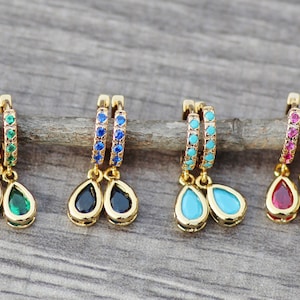 Könnte beinhalten: Vier Paar goldene Creolen mit baumelnden tropfenförmigen Edelsteinen. Die Ohrringe haben eine Reihe kleiner Edelsteine um den Reifen und einen größeren tropfenförmigen Edelstein, der unten herunterhängt. Die Edelsteine sind grün, blau, türkis und rot.