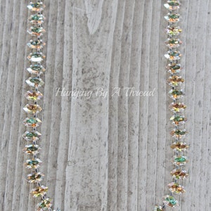 May include: A delicate necklace featuring iridescent, marquise-cut crystals. The crystals are strung together with a silver-toned chain. The necklace is displayed against a weathered wooden background. Text on the image reads "Hanging By A Thread".