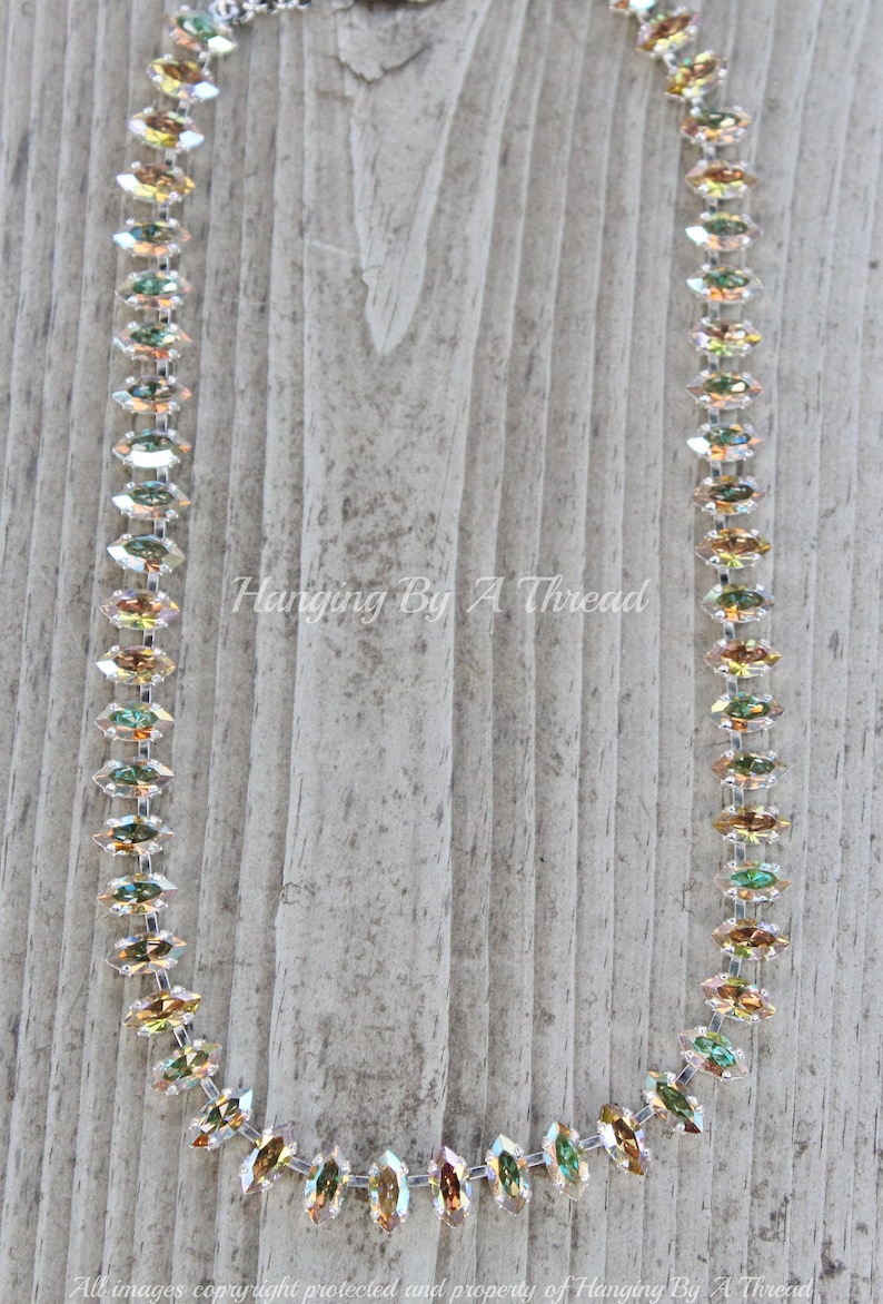 May include: A delicate necklace featuring iridescent, marquise-cut crystals. The crystals are strung together with a silver-toned chain. The necklace is displayed against a weathered wooden background. Text on the image reads "Hanging By A Thread".