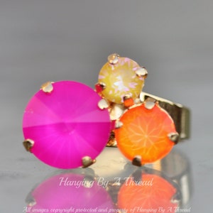 May include: A gold-toned ring featuring three large, colorful gemstones. The gemstones are a vibrant fuchsia, orange, and yellow. The ring has a cluster design with the gemstones set in a gold-colored metal.