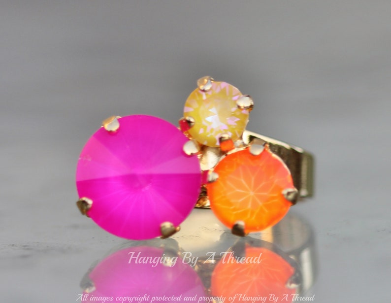 May include: A gold-toned ring featuring three large, colorful gemstones. The gemstones are a vibrant fuchsia, orange, and yellow. The ring has a cluster design with the gemstones set in a gold-colored metal.