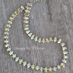 May include: A delicate necklace featuring iridescent, marquise-cut crystals set in a silver-toned chain. The crystals display a range of colors, from yellow to green. The necklace is displayed on a wooden surface. Text on the image reads "Hanging By A Thread".