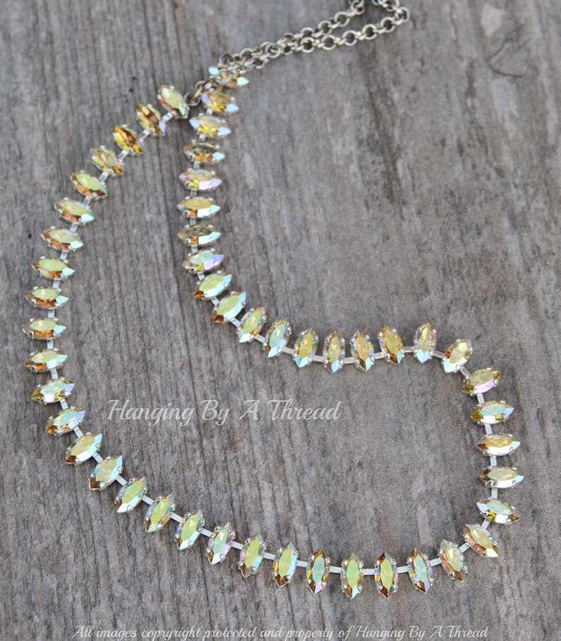May include: A delicate necklace featuring iridescent, marquise-cut crystals set in a silver-toned chain. The crystals display a range of colors, from yellow to green. The necklace is displayed on a wooden surface. Text on the image reads "Hanging By A Thread".