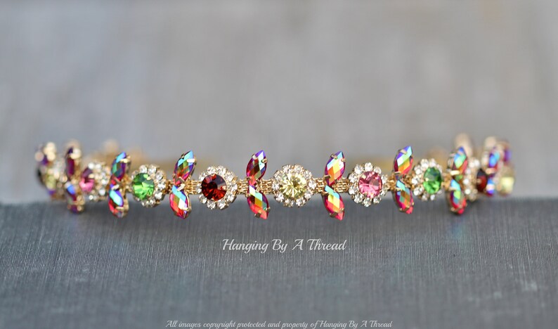 May include: A gold headband with a row of colorful iridescent rhinestones and clear crystals. The headband is decorated with small gold leaves.
