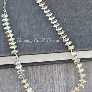May include: A silver chain necklace with iridescent, oval-shaped crystals. The crystals are set in a row, creating a sparkling effect. The necklace is displayed on a dark gray book. The text "Hanging By A Thread" is visible.
