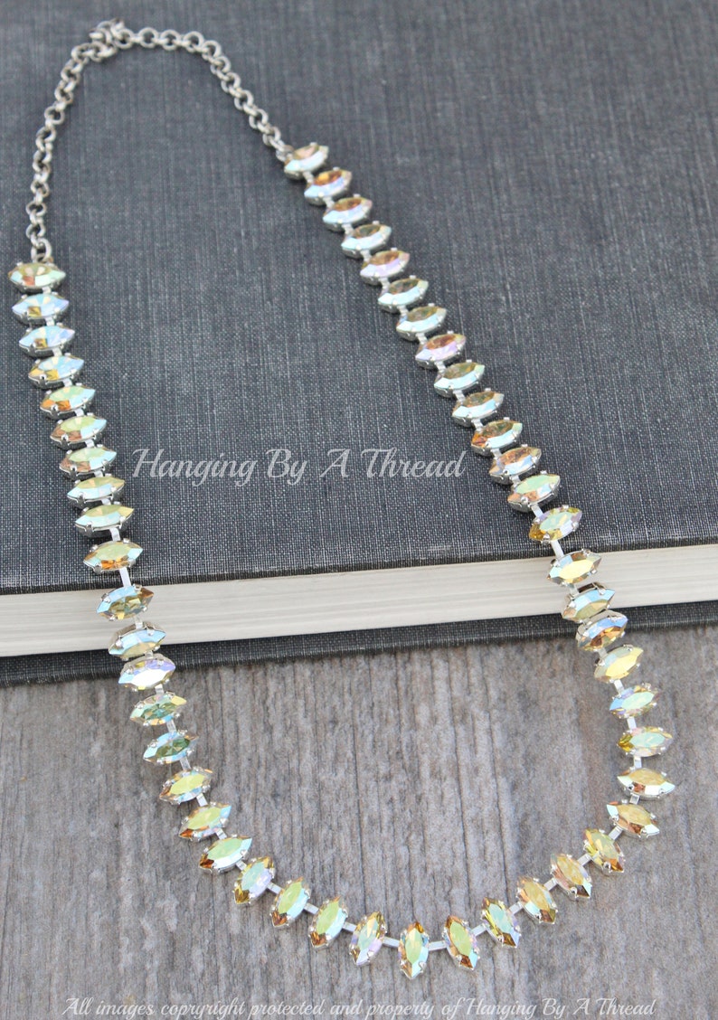 May include: A silver chain necklace with iridescent, oval-shaped crystals. The crystals are set in a row, creating a sparkling effect. The necklace is displayed on a dark gray book. The text "Hanging By A Thread" is visible.