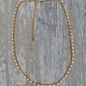 Puede incluir: Un collar de cadena dorada con peque&ntilde;os abalorios redondos colgando de &eacute;l. El collar est&aacute; sobre un fondo de madera.