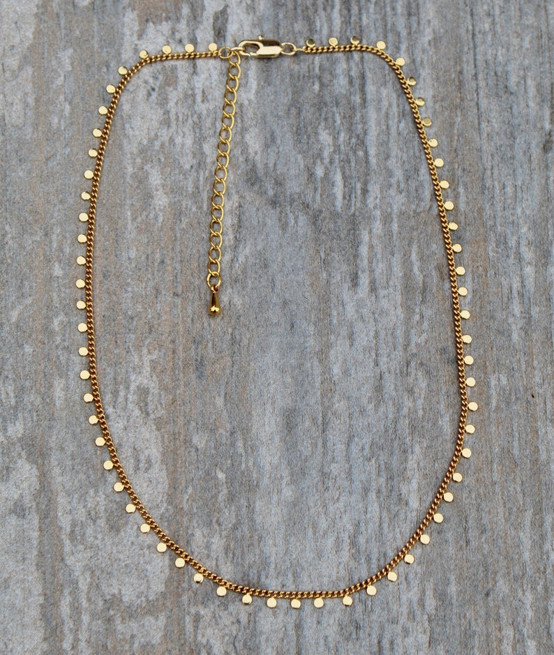 Puede incluir: Un collar de cadena dorada con peque&ntilde;os abalorios redondos colgando de &eacute;l. El collar est&aacute; sobre un fondo de madera.