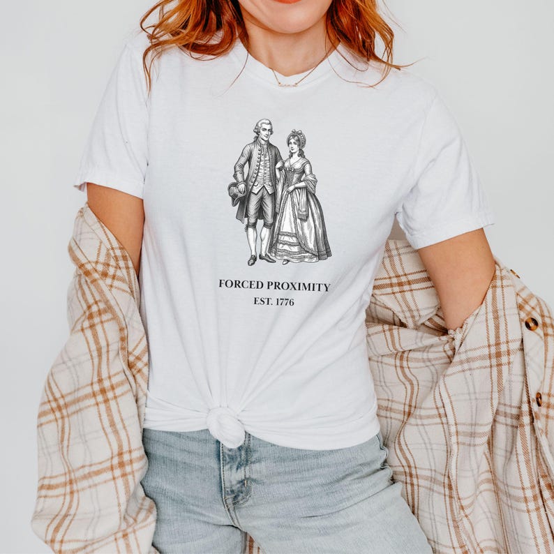 May include: White t-shirt featuring a black and white illustration of a couple in historical attire. The text "FORCED PROXIMITY EST. 1776" is printed below the image. The shirt is made of a soft material.
