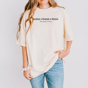 Pu&ograve; includere: T-shirt color crema con la scritta "Booktok Masktok Biketok The Holy Trinity" in nero. La maglietta ha maniche corte e una vestibilit&agrave; ampia. La persona indossa jeans blu.