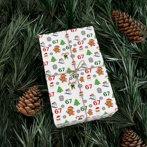May include: A wrapped gift with white wrapping paper featuring a repeating pattern of gingerbread men, Christmas trees, snowflakes, and the number "67" in red and green. The gift is tied with twine and set on a bed of pine needles and pine cones.