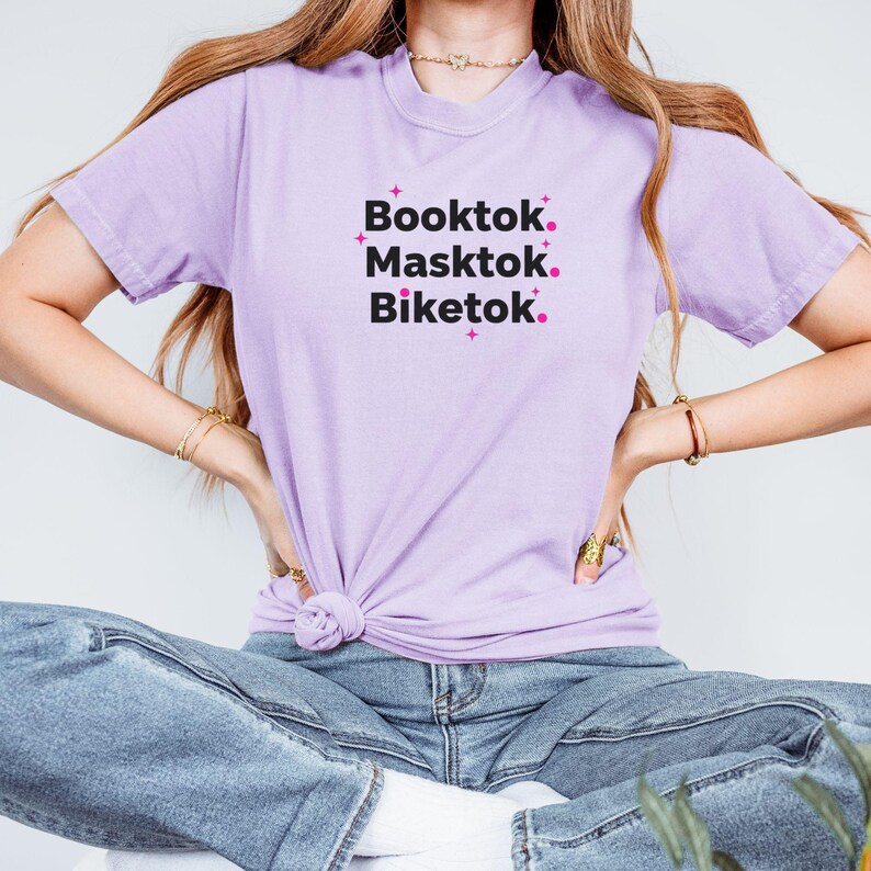 May include: A light purple t-shirt with the words "Booktok. Masktok. Biketok." in black text with pink star accents. The shirt is tied at the waist and paired with blue jeans.