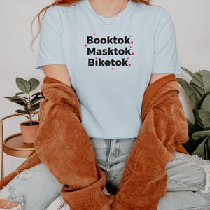 May include: A light blue t-shirt with the words "Booktok. Masktok. Biketok." in black text with pink star accents. The shirt is worn with a brown jacket and blue jeans.