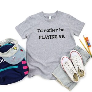 May include: A grey t-shirt with the text "I'd rather be PLAYING VR" is shown with white trainers, blue jeans, a backpack, and a visor. The trainers have red detailing. Pencils and a glue stick are also visible.