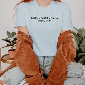Pu&ograve; includere: T-shirt azzurra con la scritta nera "Booktok &hearts; Masktok &hearts; Biketok The Holy Trinity". Sopra &egrave; indossata una giacca marrone. L'immagine &egrave; relativa alle tendenze dei social media.