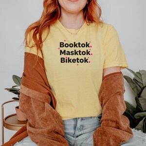 May include: A yellow t-shirt with the words "Booktok. Masktok. Biketok." in black text with pink star accents. The shirt is paired with blue jeans and a brown jacket.
