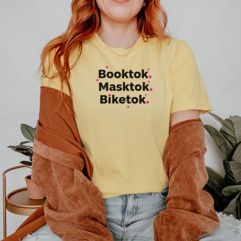 May include: A yellow t-shirt with the words "Booktok. Masktok. Biketok." in black text with pink star accents. The shirt is paired with blue jeans and a brown jacket.
