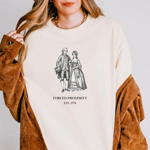 May include: Cream-colored sweatshirt featuring a black and white illustration of a couple in historical attire. The text "FORCED PROXIMITY EST. 1776" is printed below the image. The sweatshirt has long sleeves and a relaxed fit.