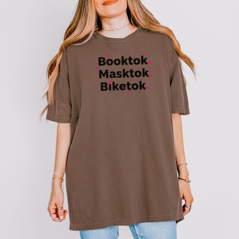 May include: Brown t-shirt with the words "Booktok, Masktok, Biketok" in black font, with pink accents. The shirt is a casual, oversized style.