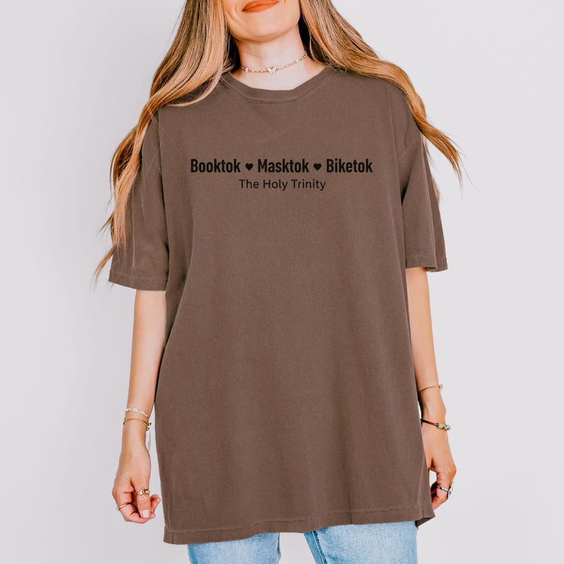 Pu&ograve; includere: Una maglietta marrone con la scritta "Booktok Masktok Biketok The Holy Trinity" in nero. La maglietta ha maniche corte e una vestibilit&agrave; ampia.