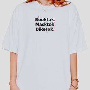 May include: White t-shirt with the words "Booktok. Masktok. Biketok." in a black font with pink accents. The shirt has a relaxed fit and short sleeves. The text is centered on the front of the shirt.