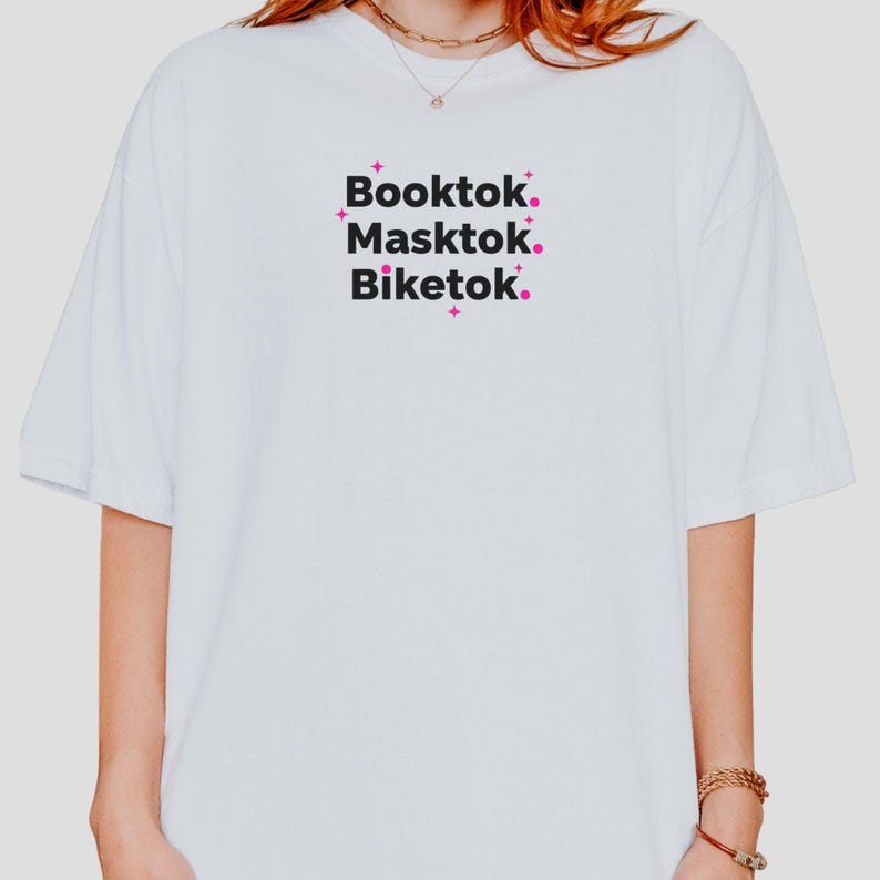 May include: White t-shirt with the words "Booktok. Masktok. Biketok." in a black font with pink accents. The shirt has a relaxed fit and short sleeves. The text is centered on the front of the shirt.