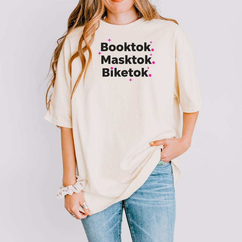 May include: Cream-colored t-shirt with the words "Booktok. Masktok. Biketok." in black text, accented with pink starbursts. The shirt is paired with blue jeans. The overall style is casual and comfortable.