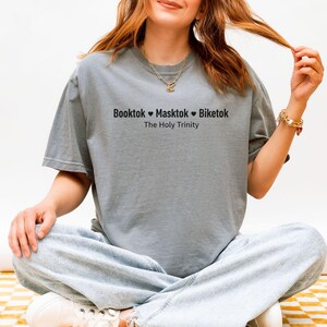 Pu&ograve; includere: Una maglietta grigia con la scritta nera "Booktok Masktok Biketok The Holy Trinity". La maglietta ha una vestibilit&agrave; ampia. La persona indossa la maglietta.