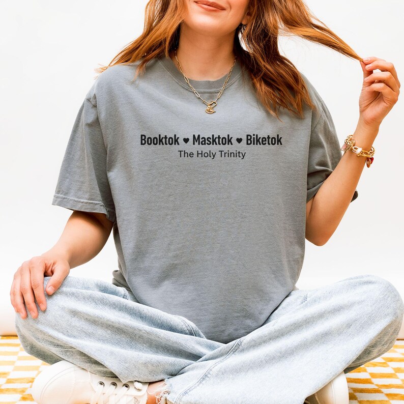 Pu&ograve; includere: Una maglietta grigia con la scritta nera "Booktok Masktok Biketok The Holy Trinity". La maglietta ha una vestibilit&agrave; ampia. La persona indossa la maglietta.