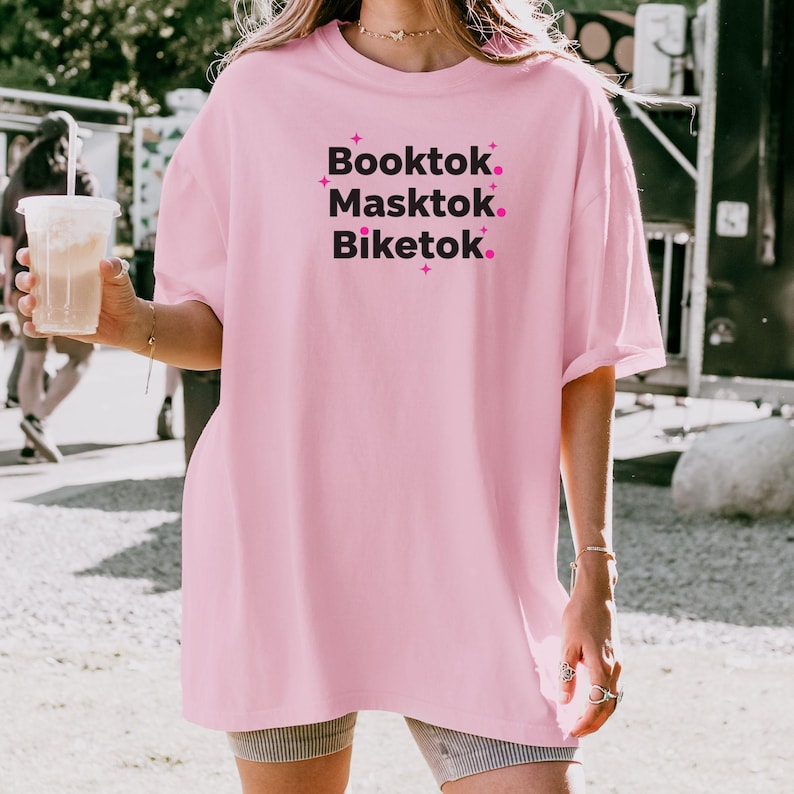 May include: A light pink oversized t-shirt with the words "Booktok. Masktok. Biketok." in black text with pink star accents. The shirt is made of a soft material and is suitable for casual wear.