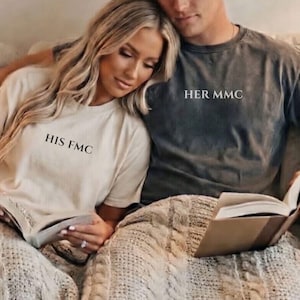 May include: Two people wearing matching t-shirts with text. One shirt is beige with "HIS FMC" printed on it, and the other is gray with "HER MMC". They are sitting together, reading a book, and snuggled under a knitted blanket.