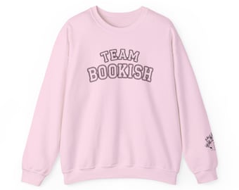 May include: A light pink sweatshirt with the text "TEAM BOOKISH" in a purple, collegiate-style font across the chest. A small floral design is on the right sleeve. The sweatshirt is a crew neck style.