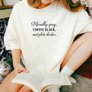 May include: Cream-colored t-shirt with the text "Morally grey, COFFEE BLACK, and plots darker" in a black, elegant font. The shirt is worn by a person holding an open book. The overall aesthetic is casual and relaxed.