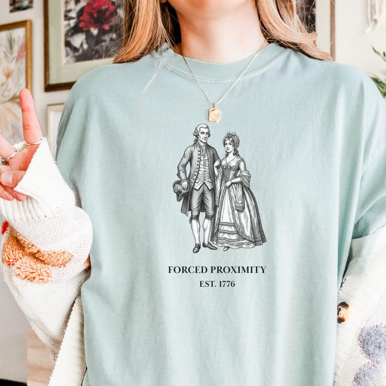 May include: A light blue t-shirt featuring a black and white illustration of a couple in historical attire. The text "FORCED PROXIMITY EST. 1776" is printed below the image. The shirt has a crew neck and short sleeves.