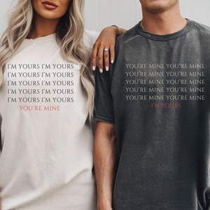 May include: Two t-shirts, one cream and one charcoal, with matching text. The cream shirt reads "I'M YOURS" repeated, with "YOU'RE MINE" in red. The charcoal shirt reads "YOU'RE MINE" repeated, with "I'M YOURS" in red.