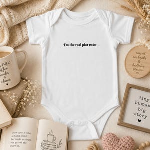 May include: A white short-sleeved baby bodysuit featuring the black printed text I'm the real plot twist centered on the chest. The garment has a round neckline and a snap-button closure at the bottom, designed for infants with a simple, clean aesthetic.