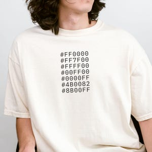 May include: Cream-colored t-shirt with a black graphic of color hex codes. The codes are arranged vertically, showcasing a range of colors. The shirt has a relaxed fit and short sleeves, suitable for casual wear.